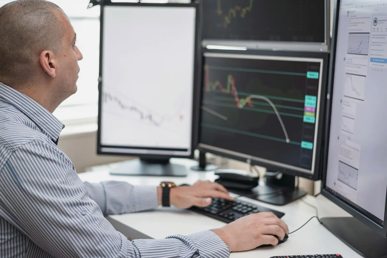 An analyst at a desk studies charts across several monitors by a bright window.