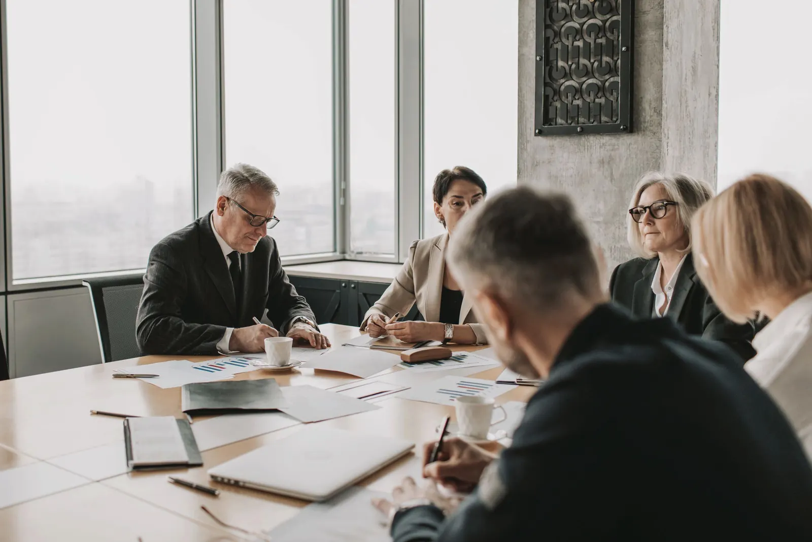 Board members take notes on papers spread across a long table in a bright, high-floor room.