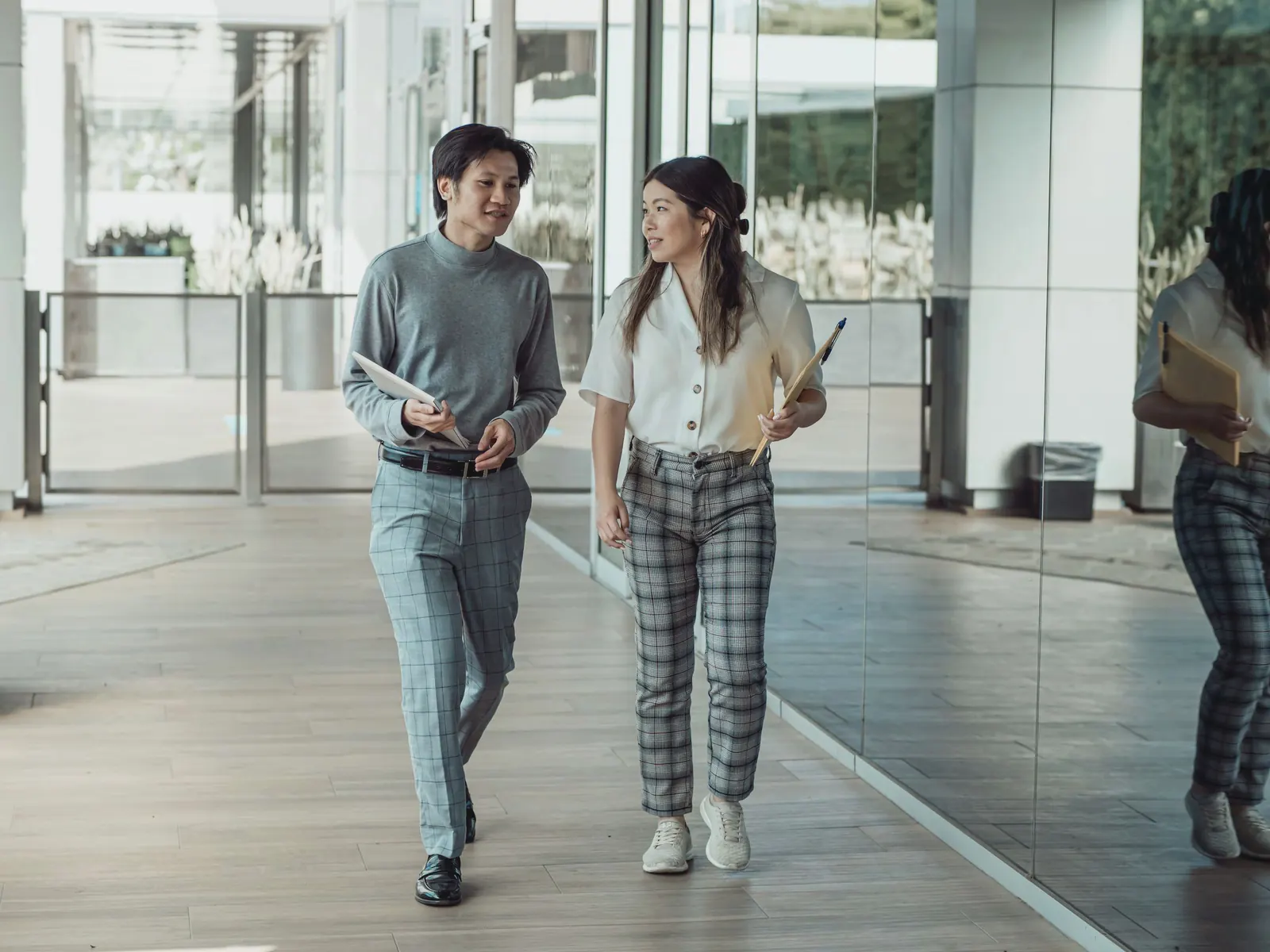 Two colleagues carrying folders talk as they walk down a bright glass corridor.