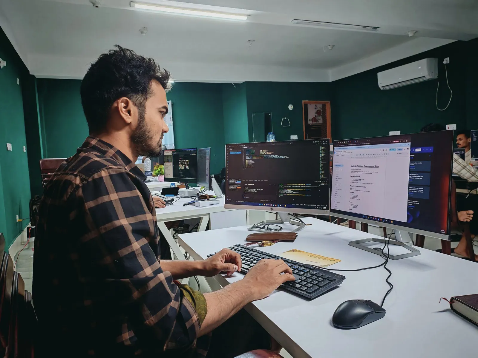 A developer types at a desk with two monitors showing code in an office with deep green walls.