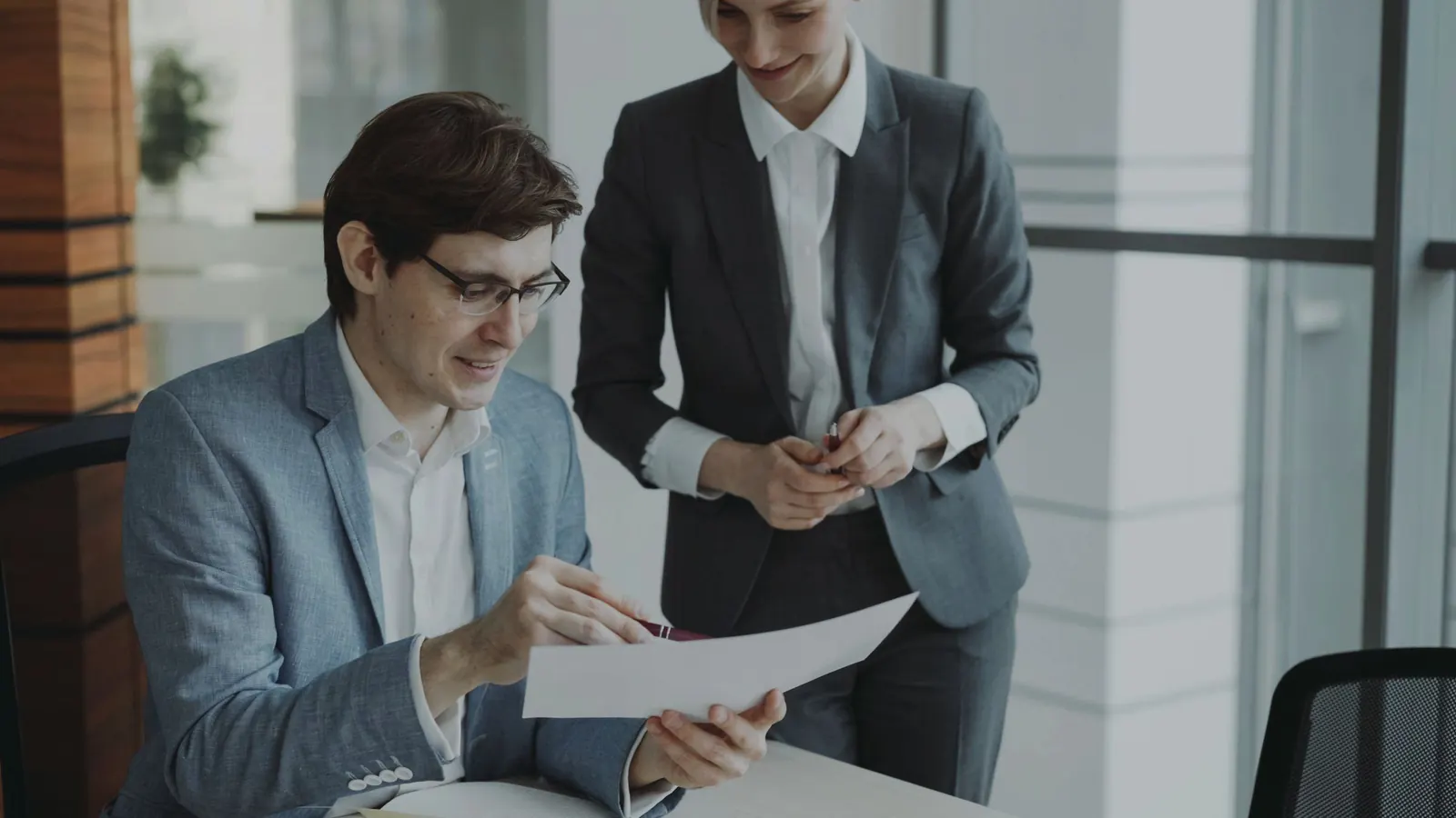 A colleague stands beside a seated partner as they review a printed page together.