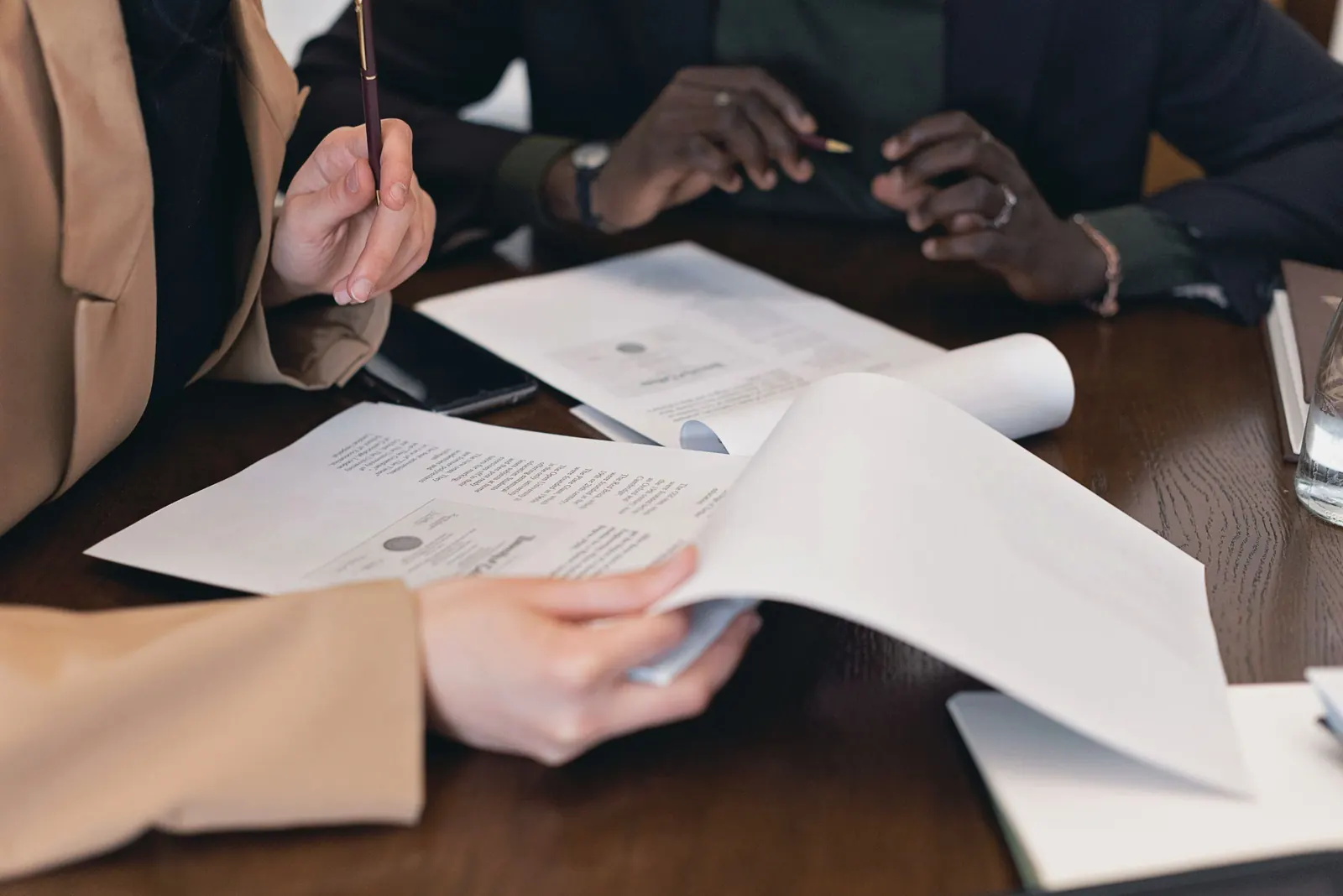 Close-up of hands with pens over printed documents on a dark wooden meeting table.