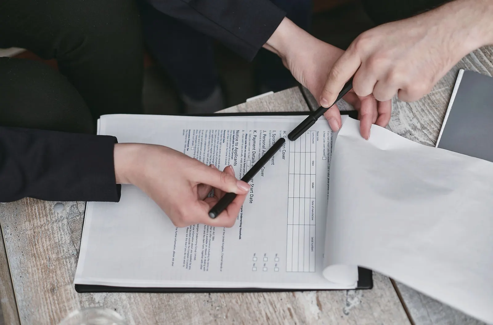 Two people point with pens at a form on a clipboard laid on a weathered table.
