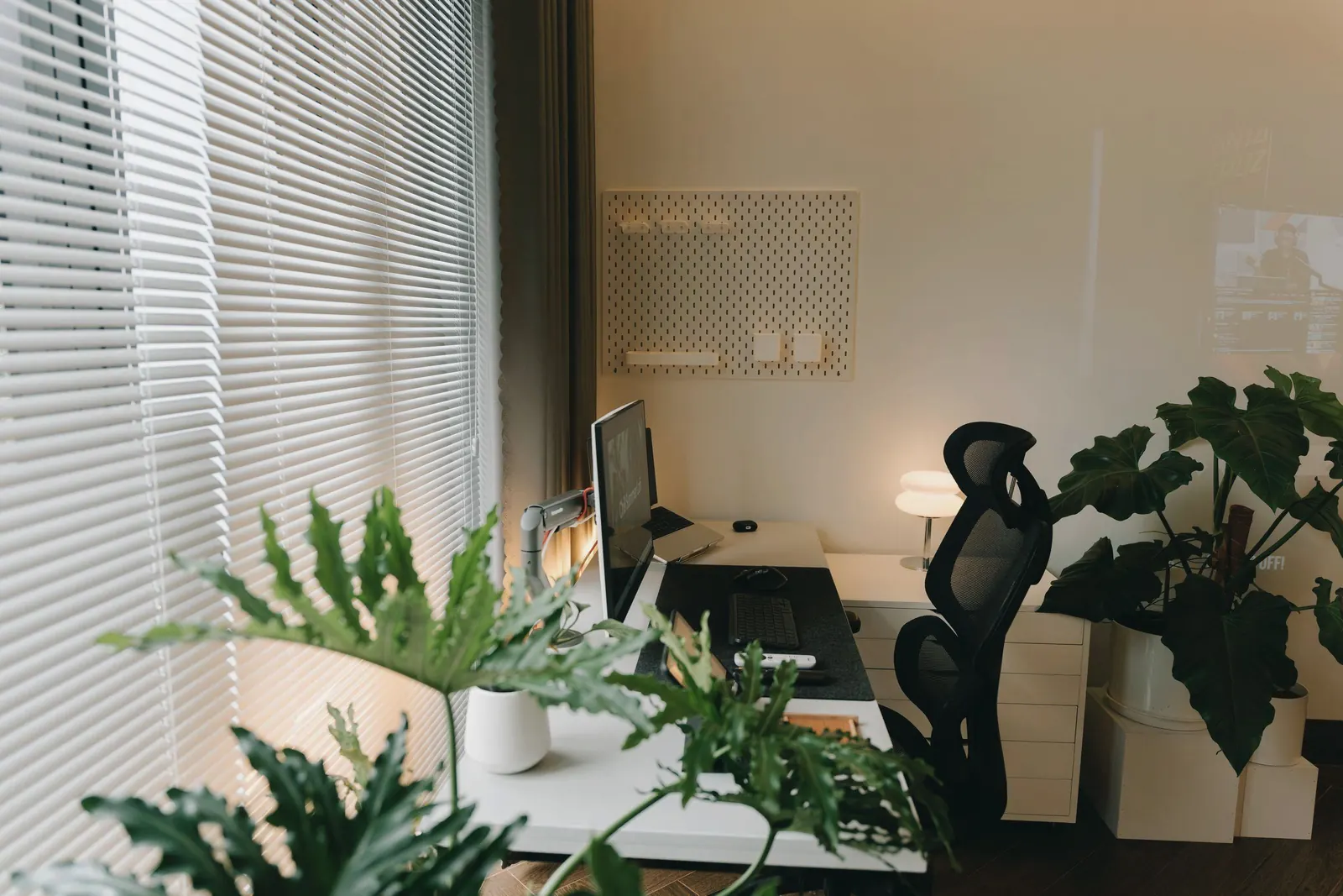 A home desk with a monitor and leafy plants beside window blinds letting in daylight.
