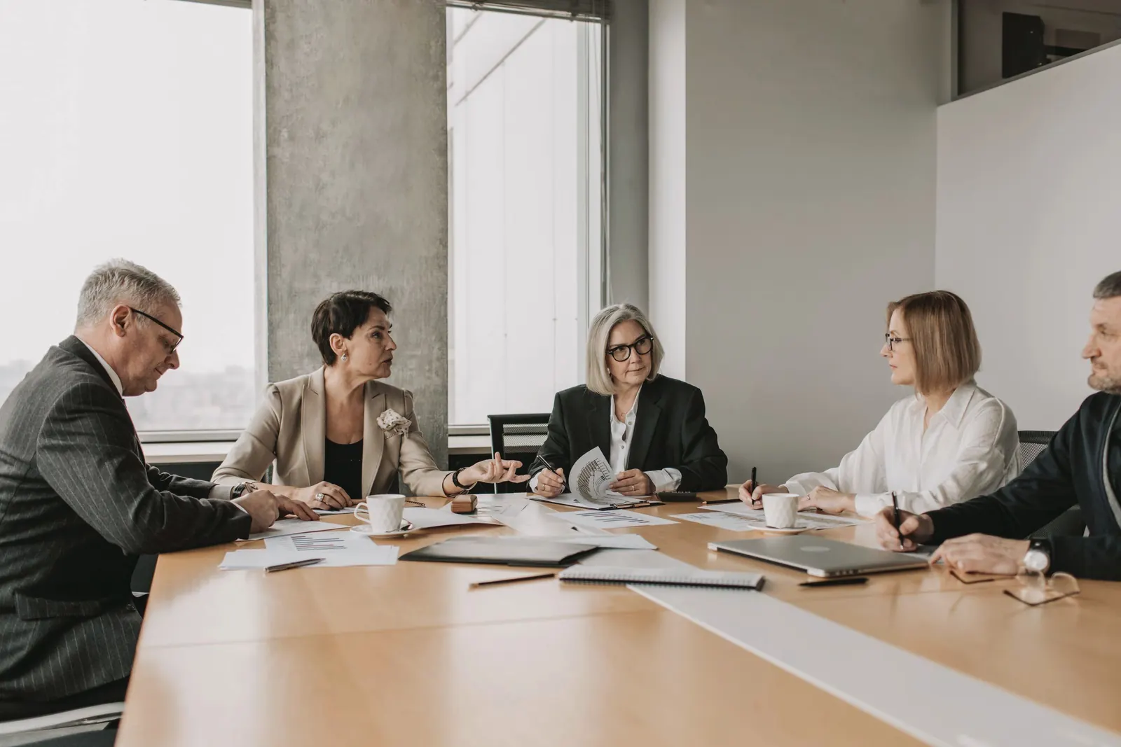 Five senior colleagues talk around a light wooden conference table beside tall windows.