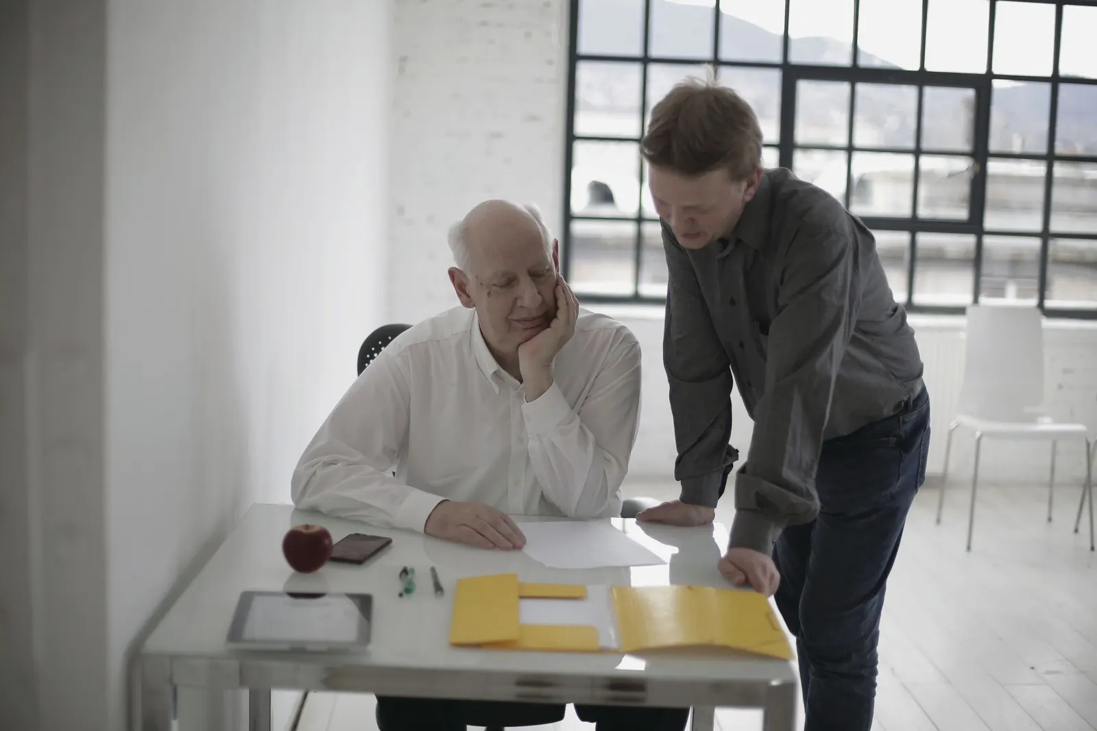 A younger colleague leans over a desk to talk through papers with a senior colleague in a white loft office.