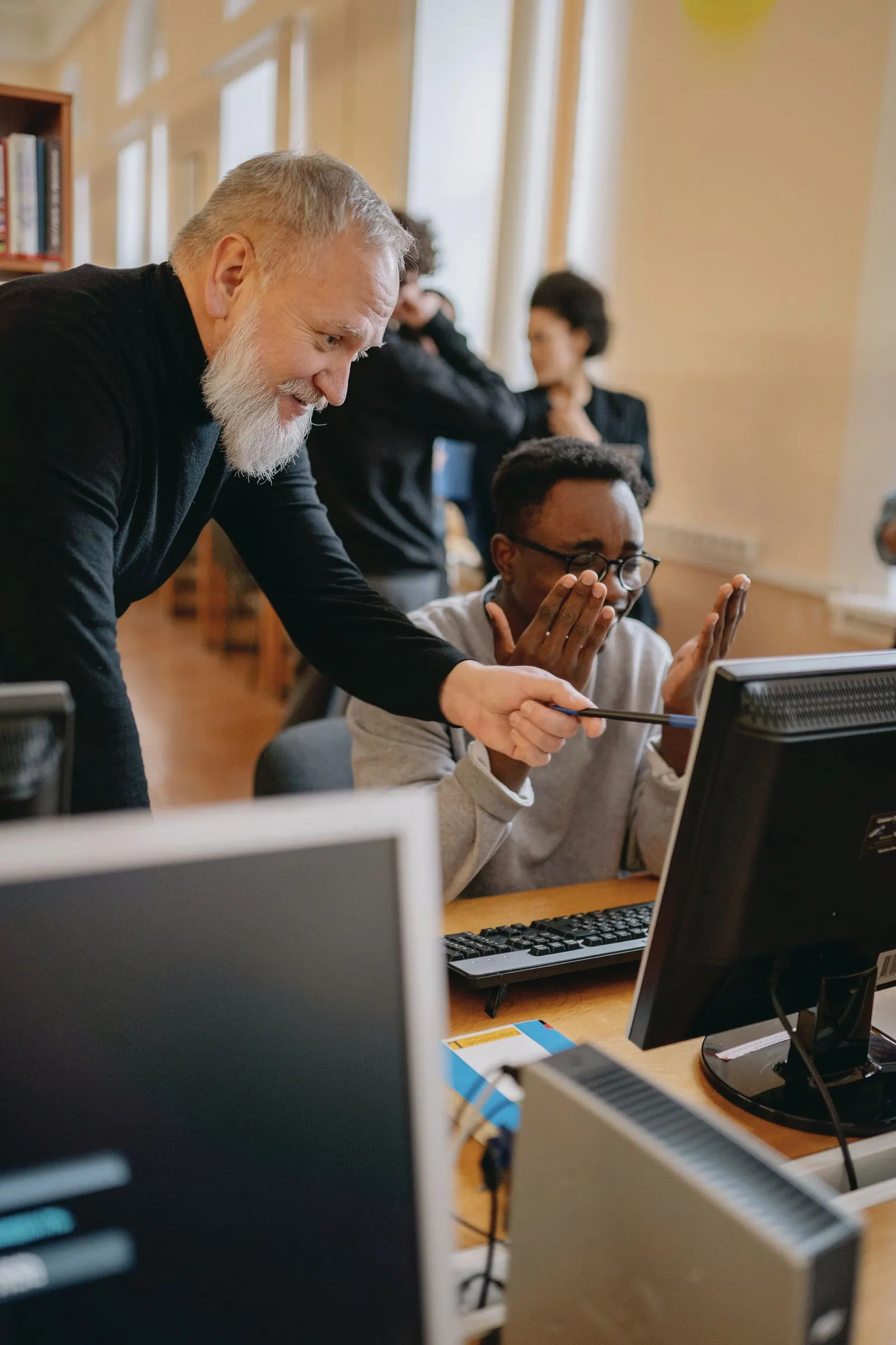 An older mentor leans in and points at a screen as a younger colleague reacts at his computer.