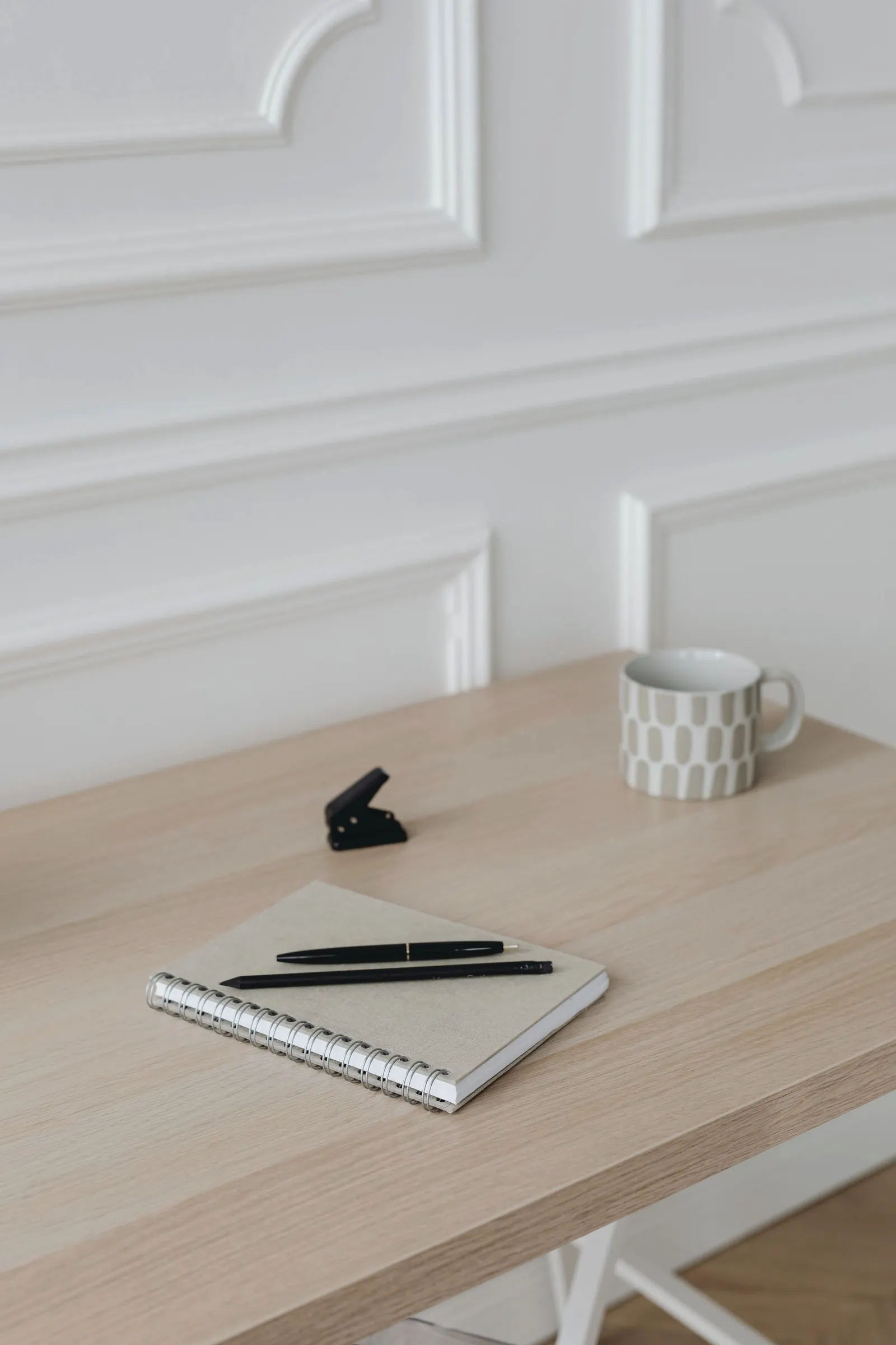 A closed notebook with two pens and a ceramic cup on a pale wooden desk against a white wall.