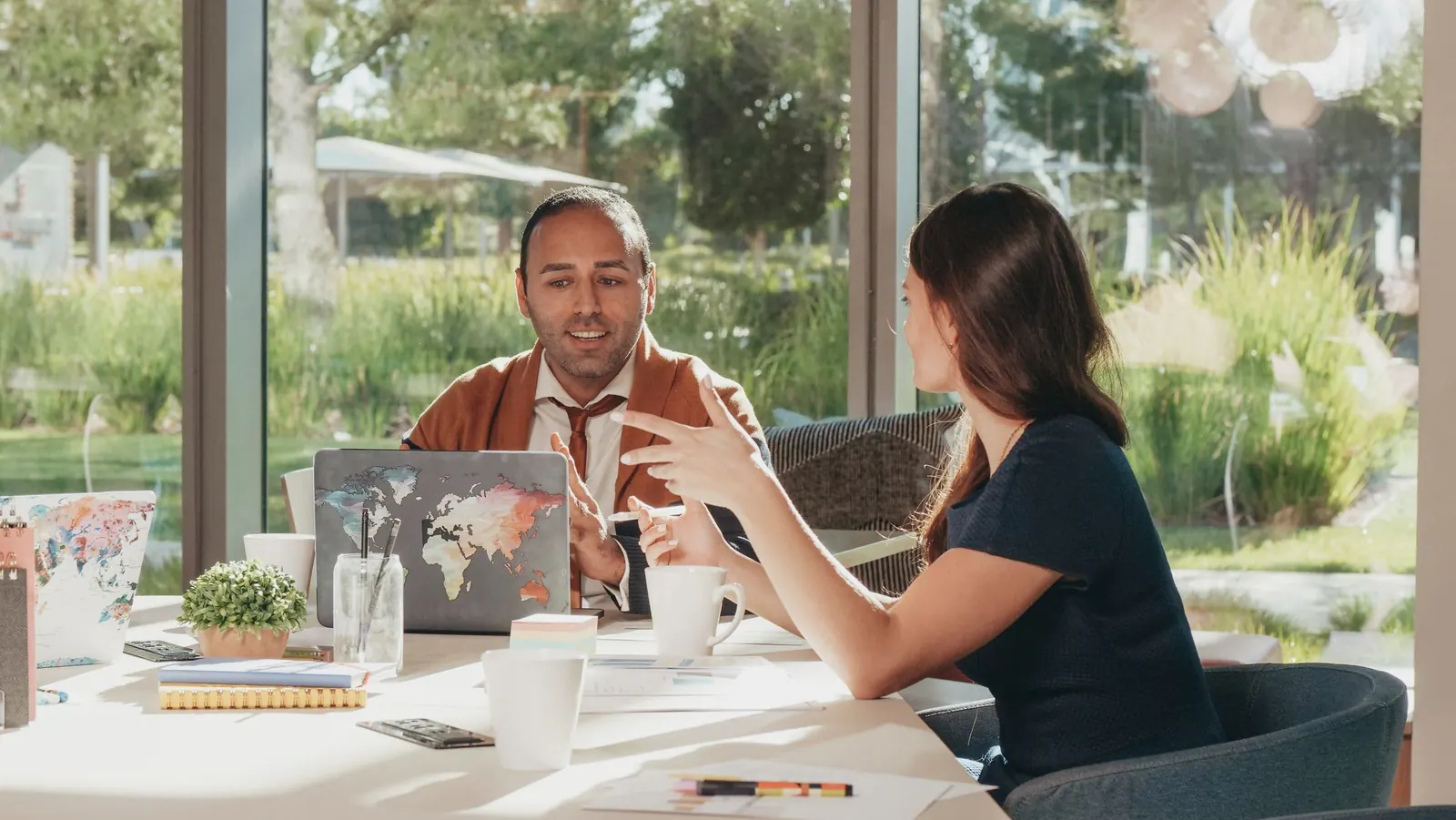 Two partners talk across a table by a sunny window that looks onto a garden.