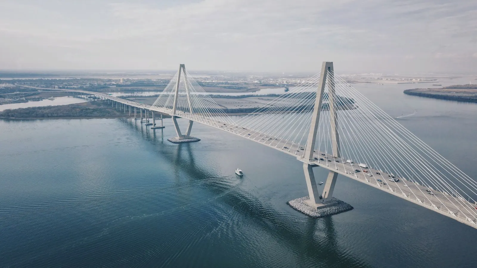 Aerial view of the Ravenel Bridge spanning still water toward low marsh islands under a hazy sky.
