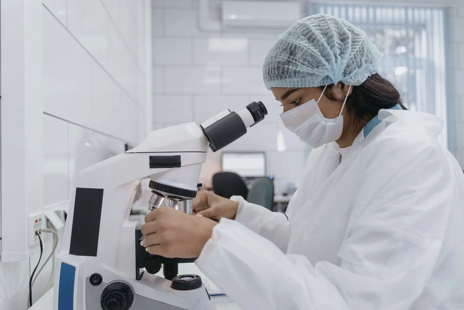 A scientist in a lab coat and cap studies a sample through a microscope at a white bench.