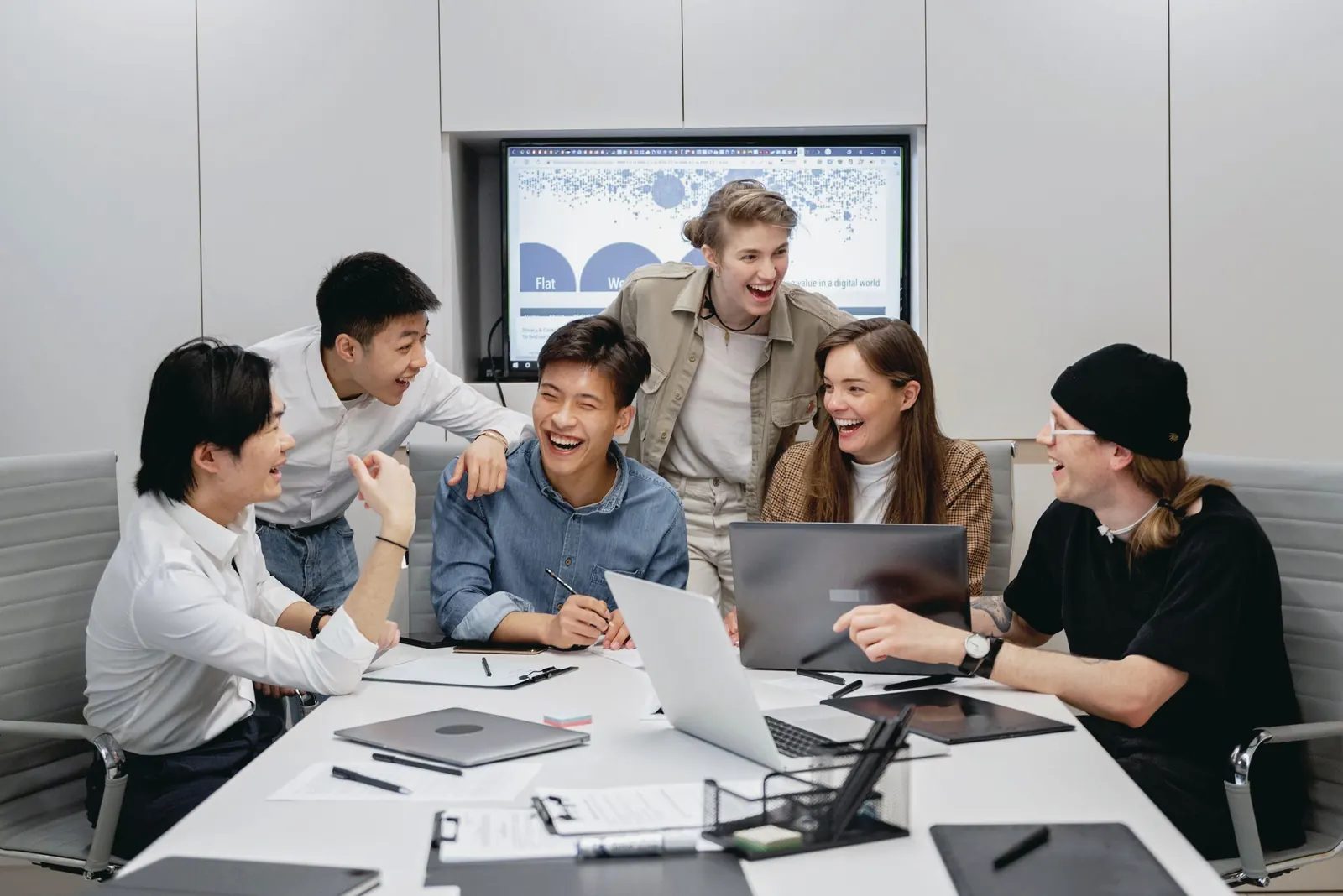 A small team laughs together around a white meeting table with laptops and notes.