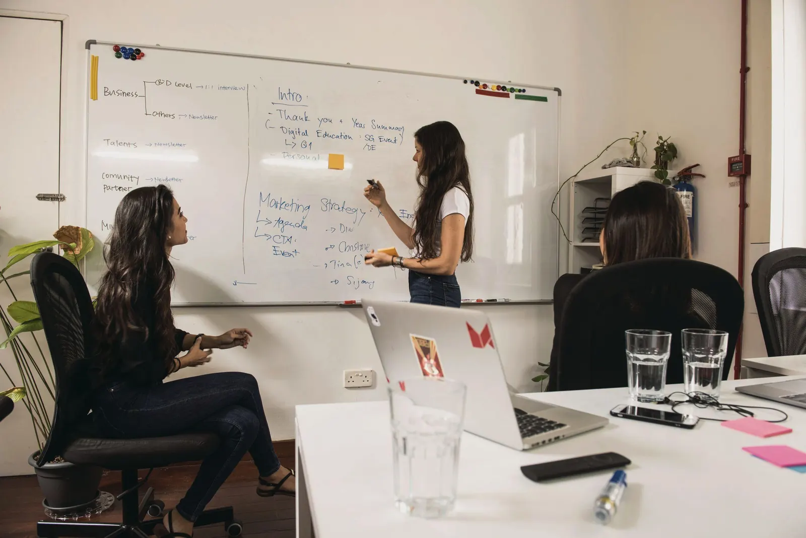 A consultant writes a plan on a whiteboard while colleagues at the table watch and discuss.