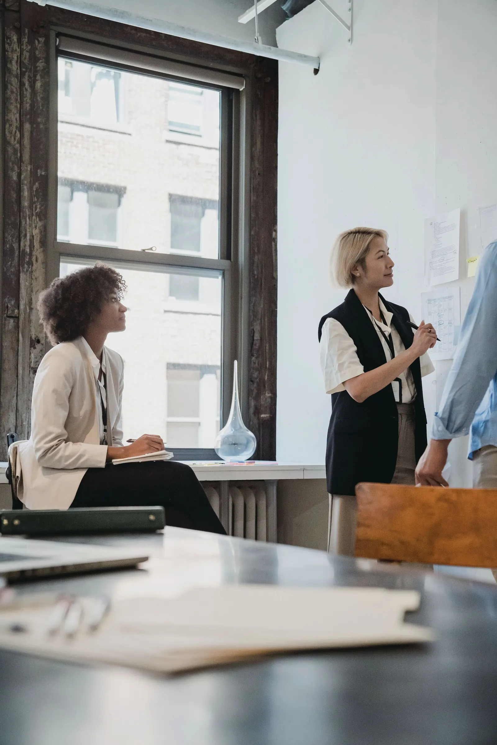 Two colleagues in a bright loft office listen as a teammate presents at the wall.
